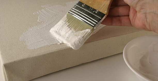 Closeup of a woman's hand painting gesso primer onto a stretched canvas to seal it